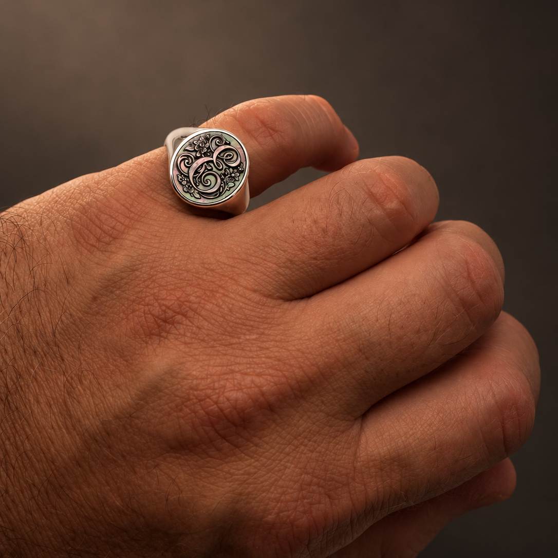Mano maschile che indossa un anello con sigillo, decorato con il monogramma della lettera E in corsivo e motivi floreali incisi su madreperla grigia iridescente. - Hand wearing a gold signet ring featuring a cursive letter E monogram and floral motifs engraved on iridescent grey mother-of-pearl.