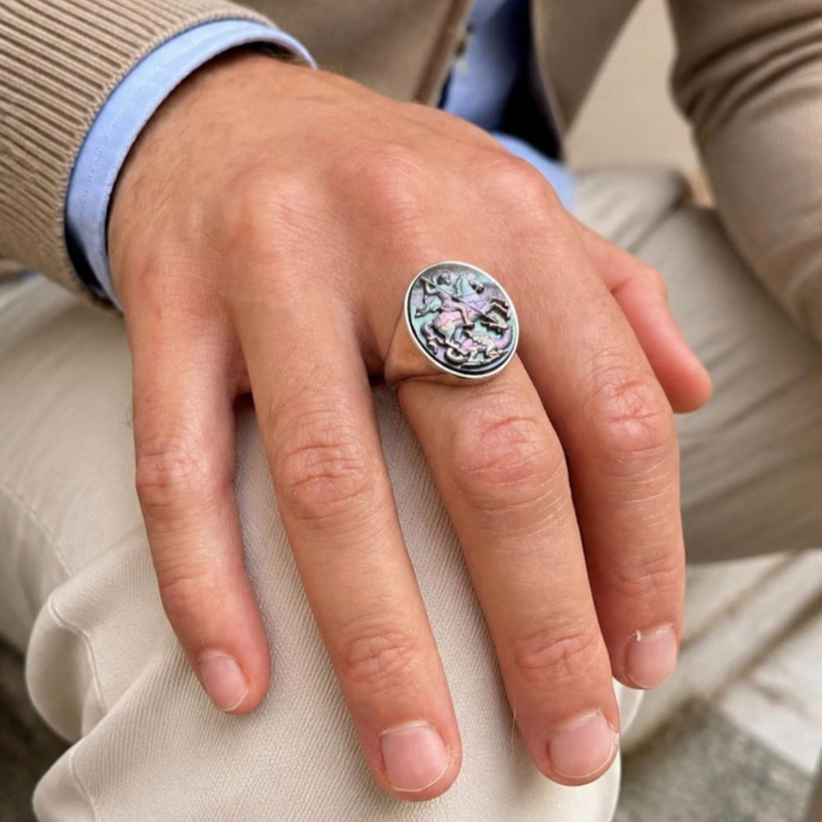 Dettaglio della mano di un uomo appoggiata sul ginocchio, che indossa un anello sigillo in argento con cammeo in madreperla scura iridescente raffigurante San Giorgio e il drago - Close-up of a man's hand resting on his knee, wearing a silver signet ring with a dark iridescent mother-of-pearl cameo depicting Saint George slaying the dragon.